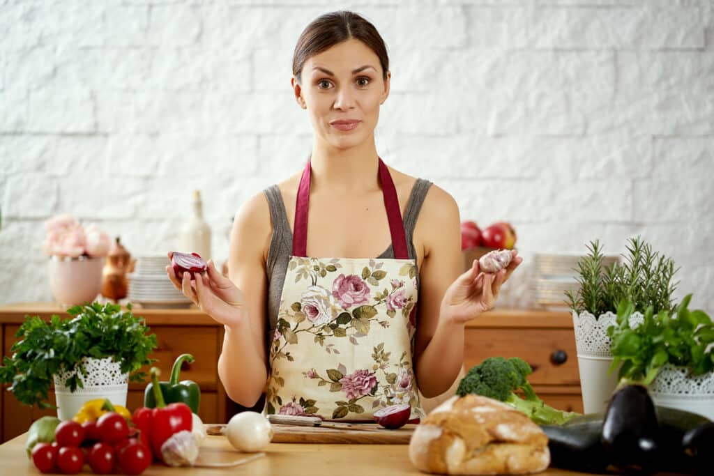 Greek-American woman in a floral apron holding onion and garlic at a kitchen counter with fresh vegetables