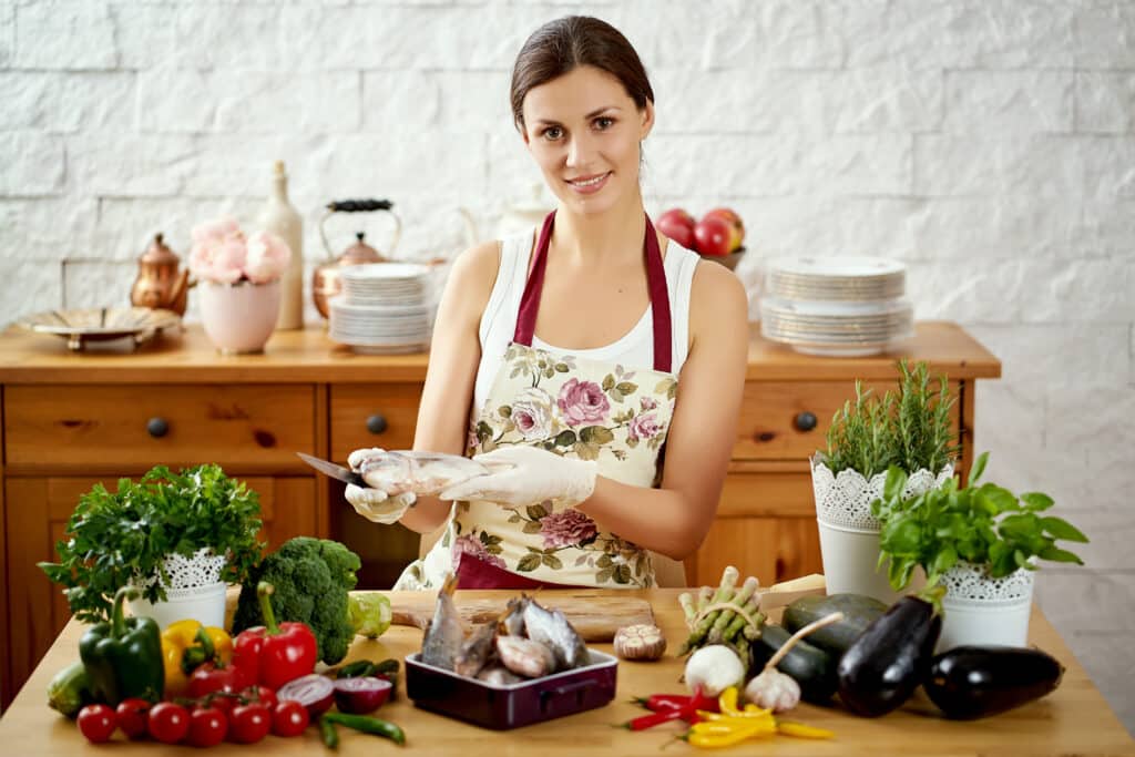 Woman cleaning fresh fish at a wooden kitchen table surrounded by herbs and vegetables