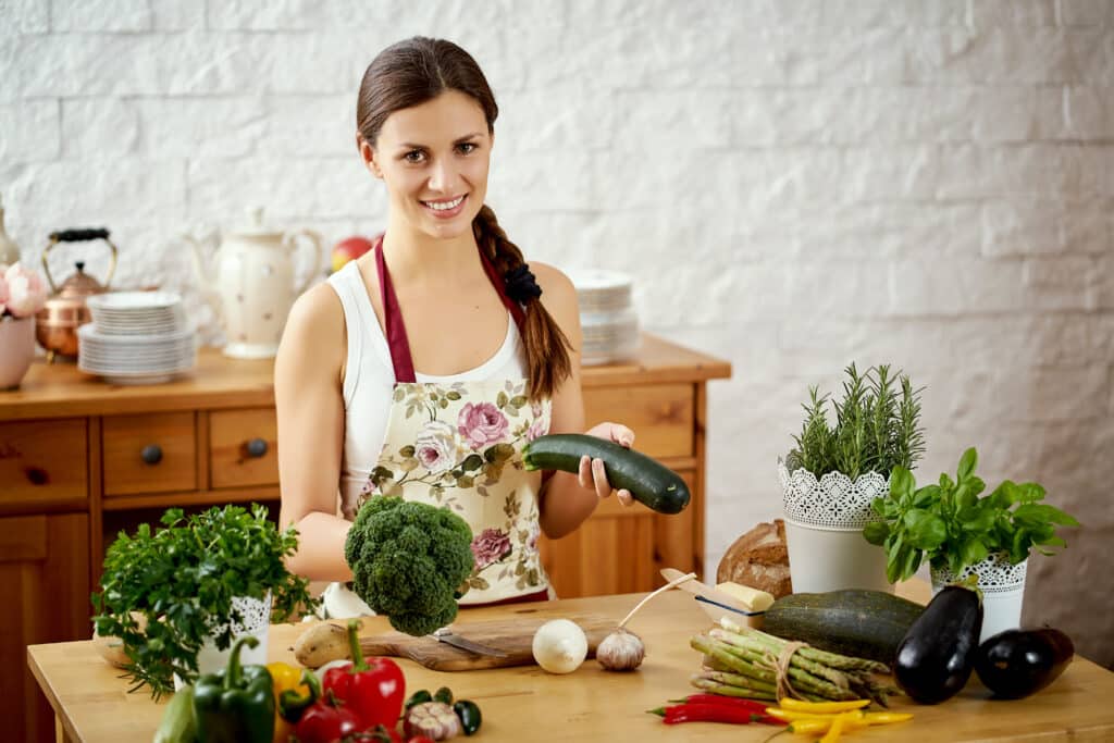 Smiling woman in floral apron holding zucchini and broccoli at a kitchen counter with fresh vegetables